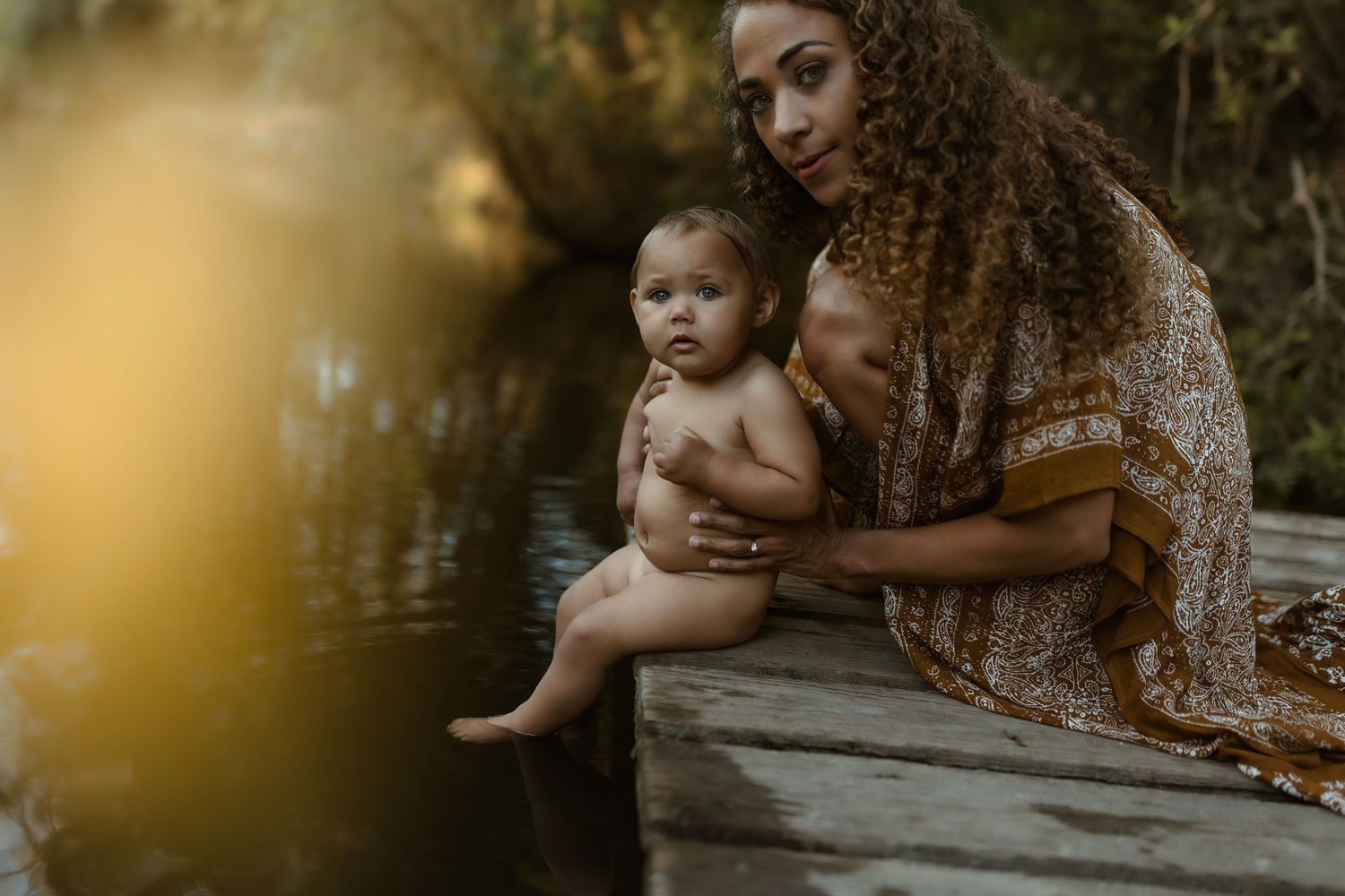 Curly-haired mother sits on wooden dock holding baby near reflective forest lake at dusk.