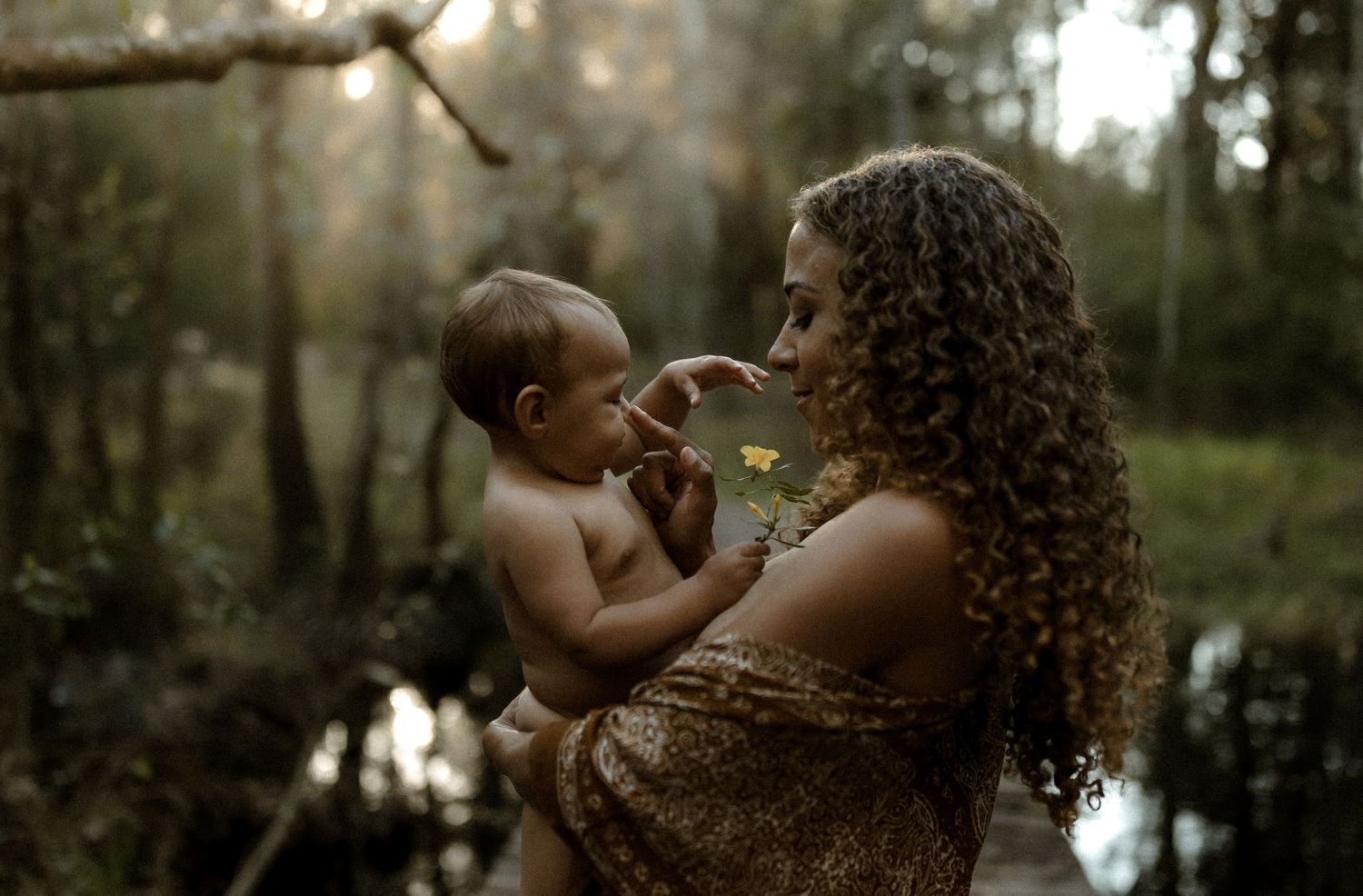 A curly-haired mother smiles at her baby while offering a small yellow flower in a forest setting.