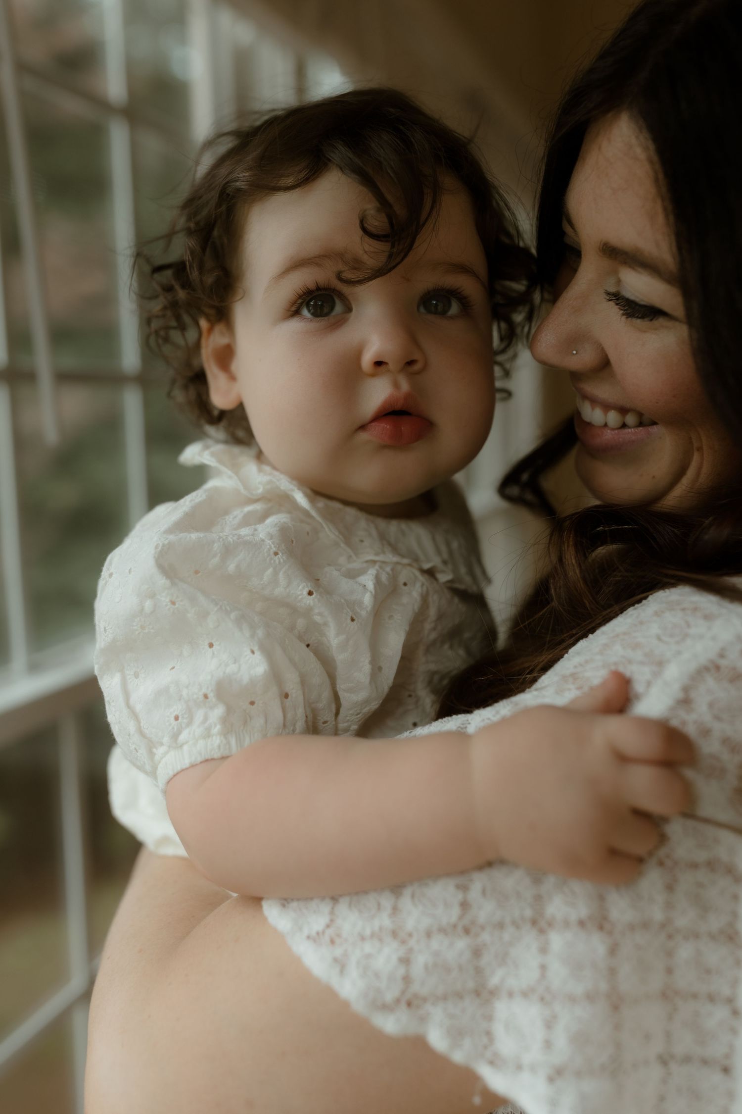 A smiling mother holds her curly-haired baby in a white dress near a sunlit window.