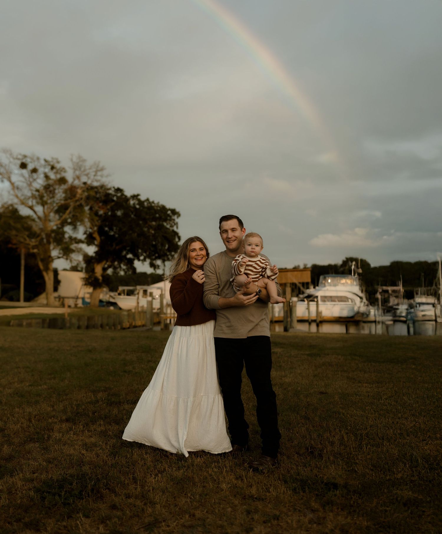 A happy family of three poses under a rainbow near a marina, with a woman in a white skirt, man, and baby.