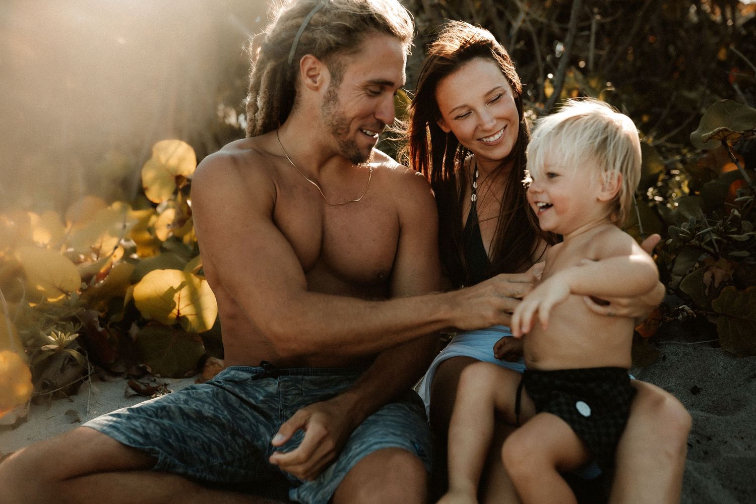 Happy family with parents and toddler laughing together on a sunny beach in warm golden light.