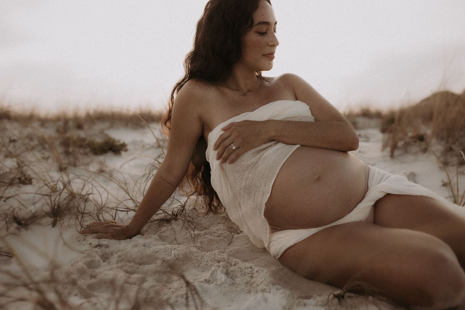 Pregnant woman draped in white fabric reclines gracefully on sandy beach dunes during a serene maternity photoshoot.