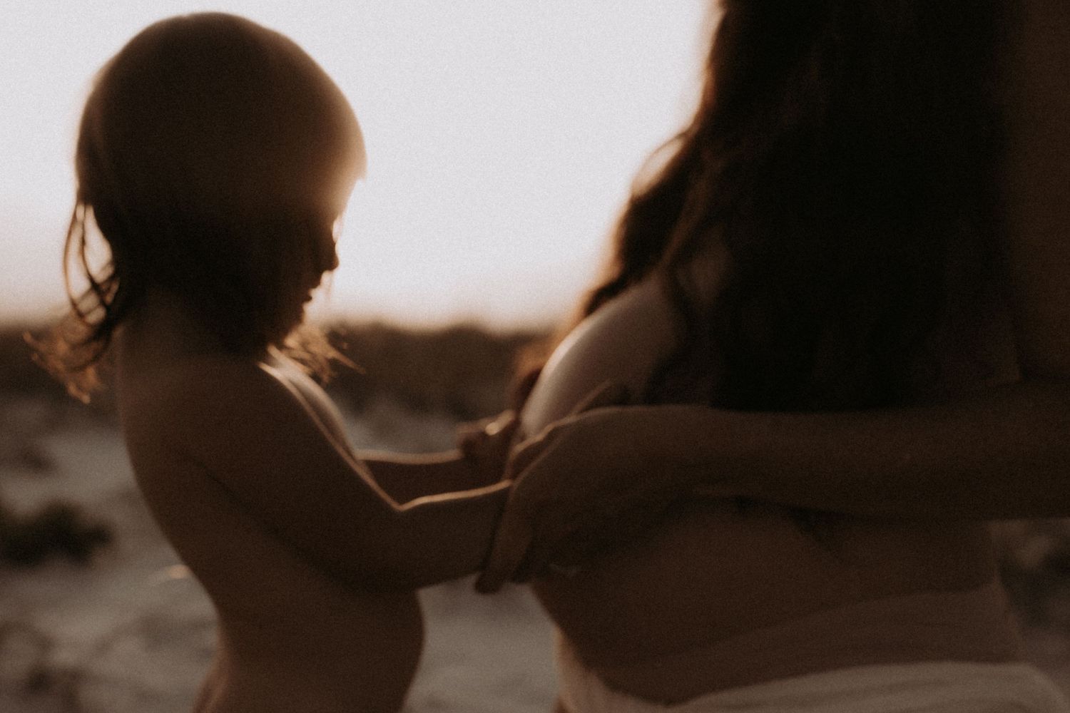 Silhouette of toddler touching pregnant mothers belly in warm golden backlit beach setting.