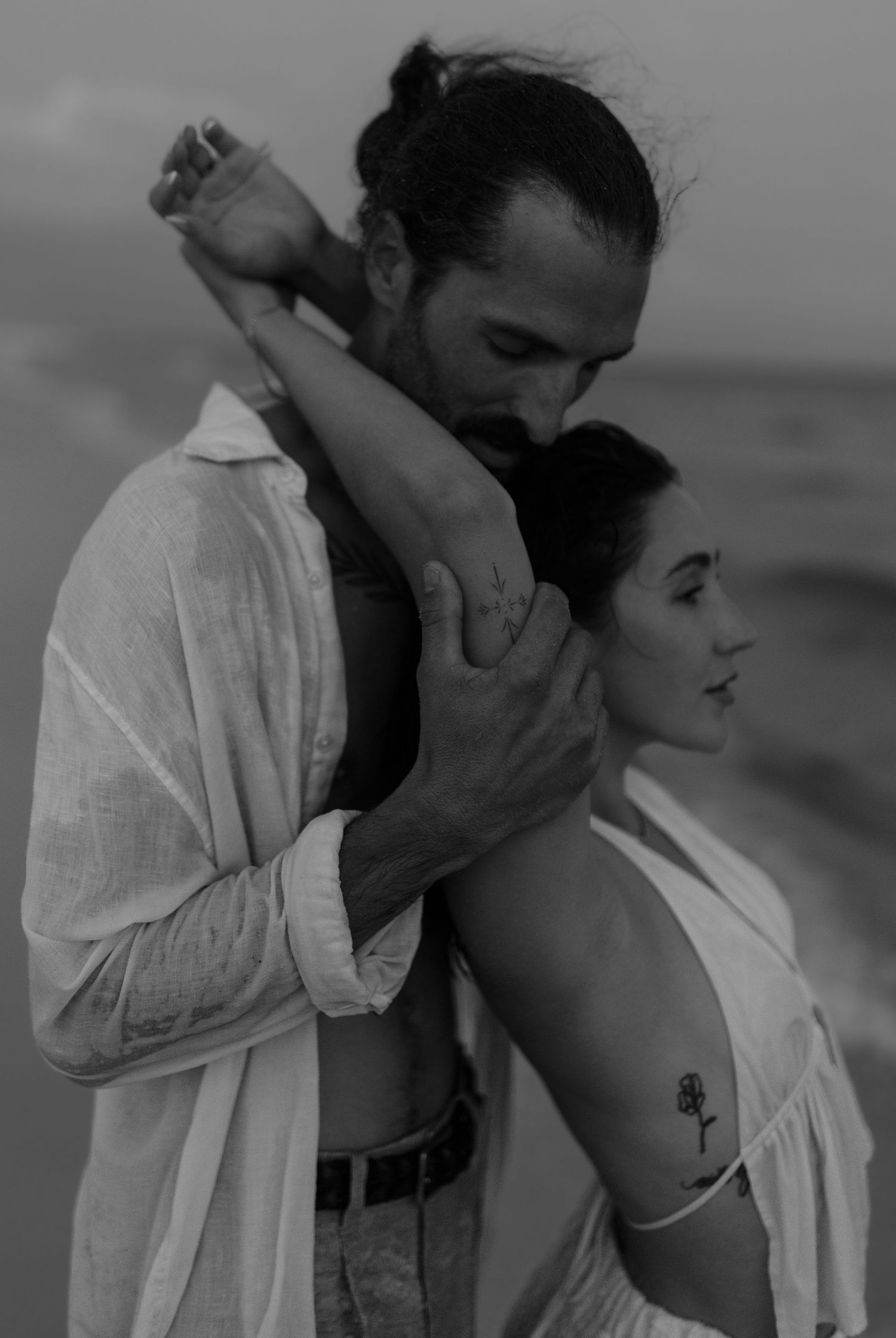 Black and white photo of a man embracing a woman from behind on a beach.