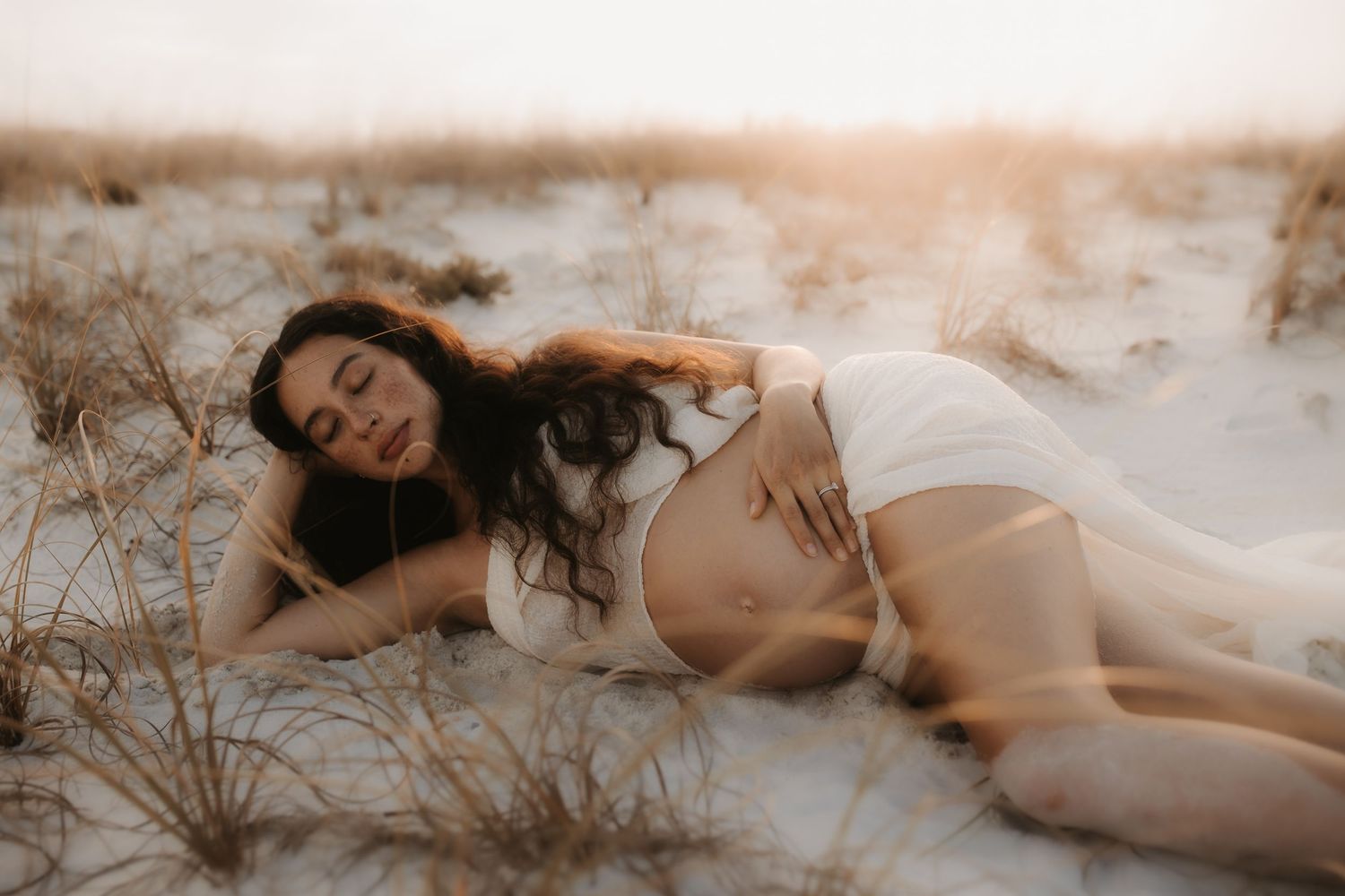 Pregnant woman in white lingerie lying on sandy beach dunes at golden sunset hour.