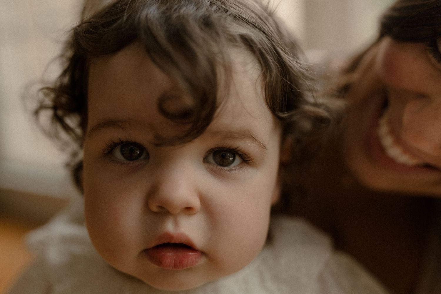 Close-up portrait of a curious baby with dark curly hair and bright eyes, with a smiling adult in the background.