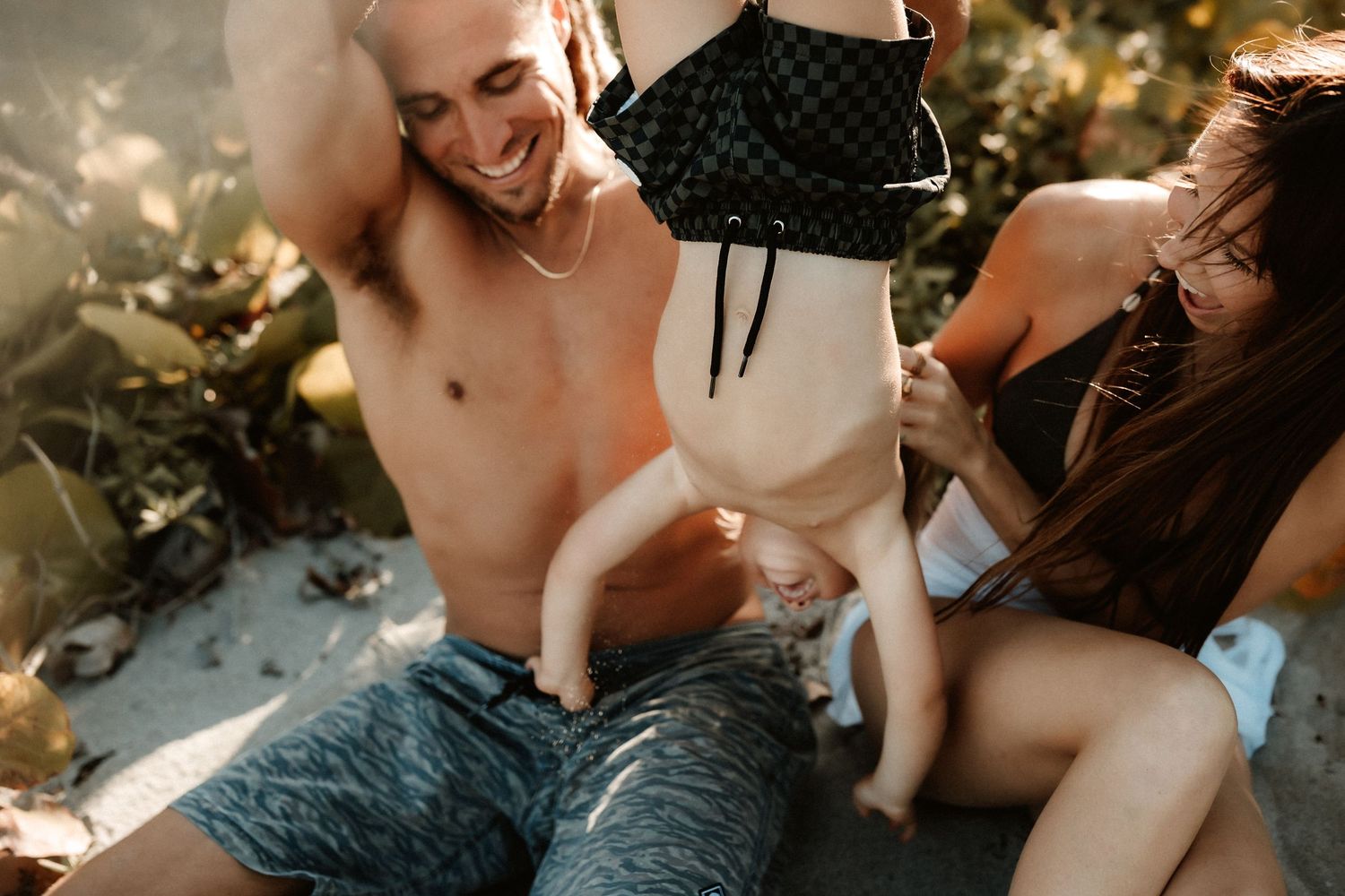 Shirtless man and woman playfully holding a baby upside down outdoors in warm sunlight.