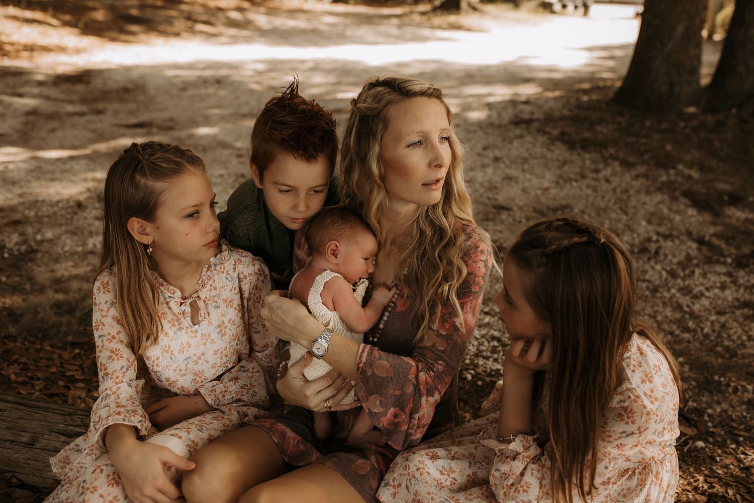 A mother sits outdoors holding a newborn baby surrounded by three young children in floral outfits under shaded trees.