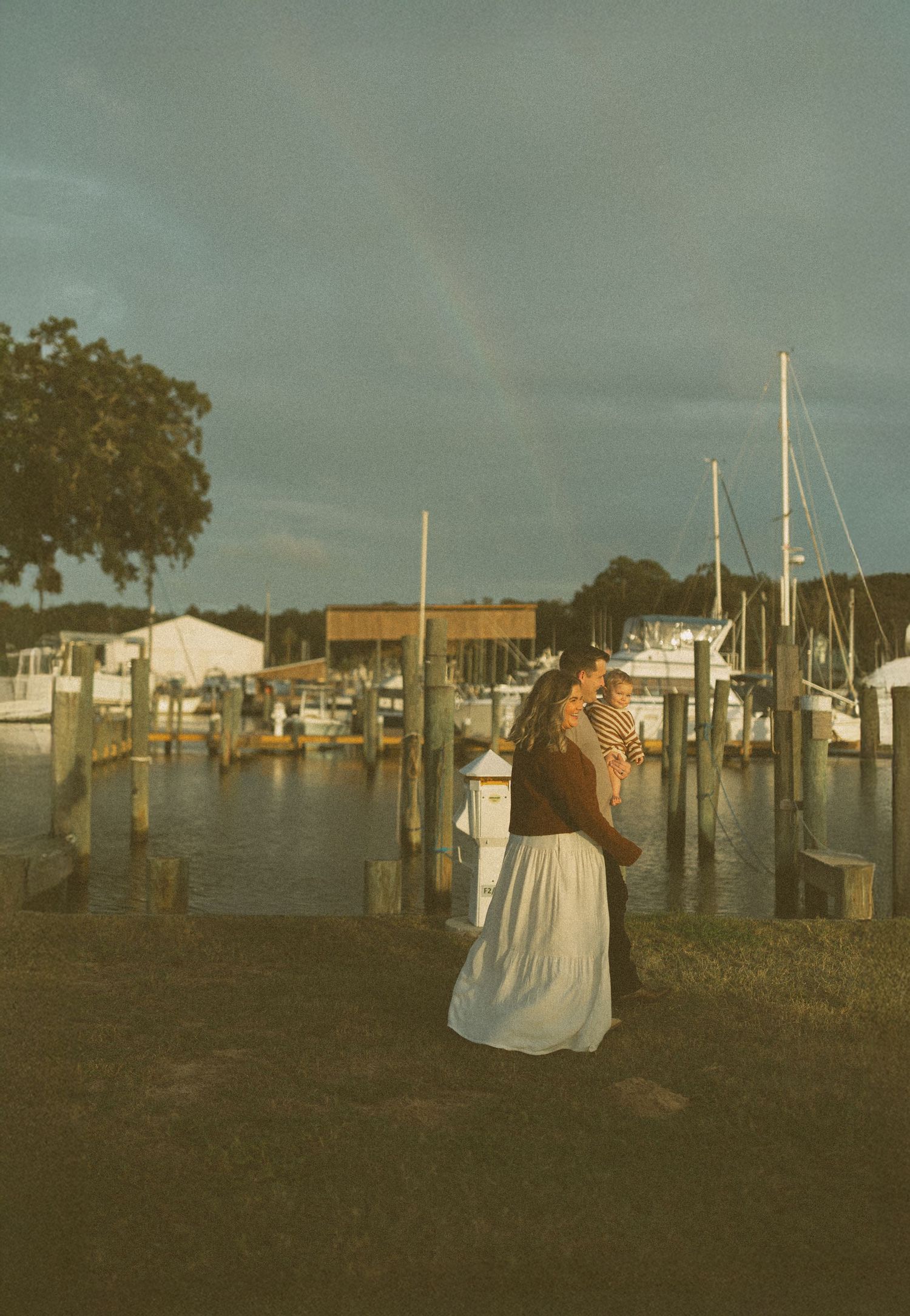 A woman in a white skirt holds a baby at a marina dock with sailboats and a rainbow in the cloudy sky.