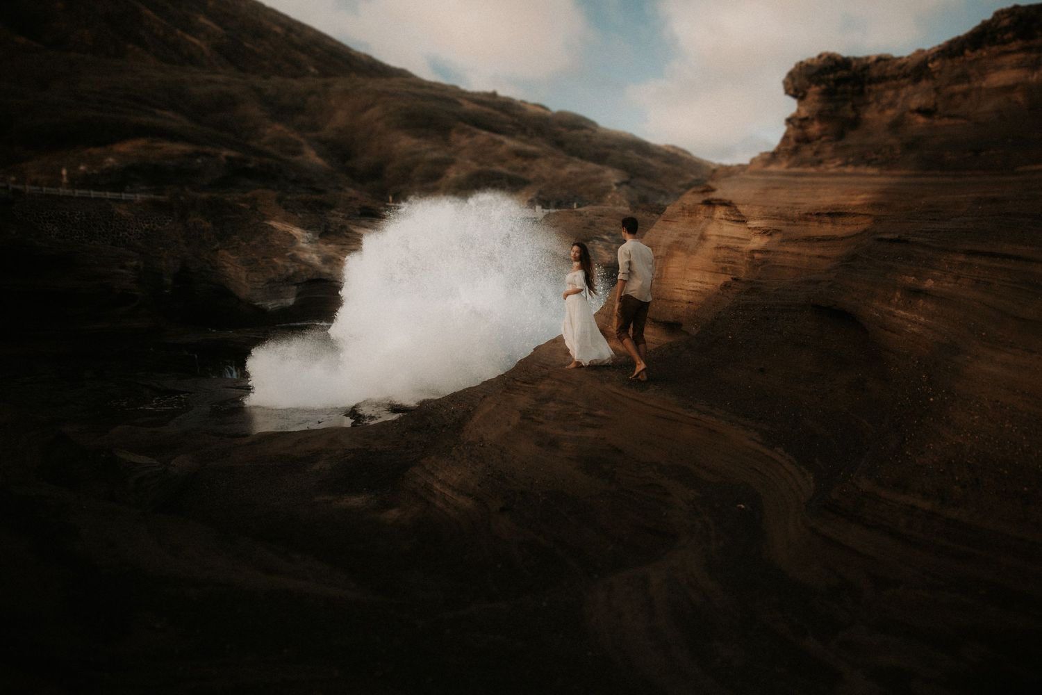 Couple standing on dark volcanic rocks with a dramatic crashing wave behind them during an adventurous elopement shoot.