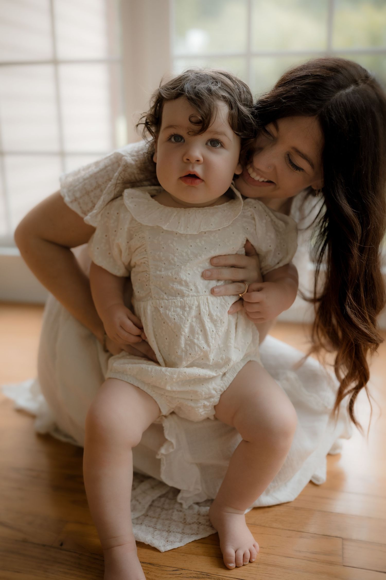 A smiling mother in a white dress holds her curly-haired baby in a cream romper on a wooden floor.