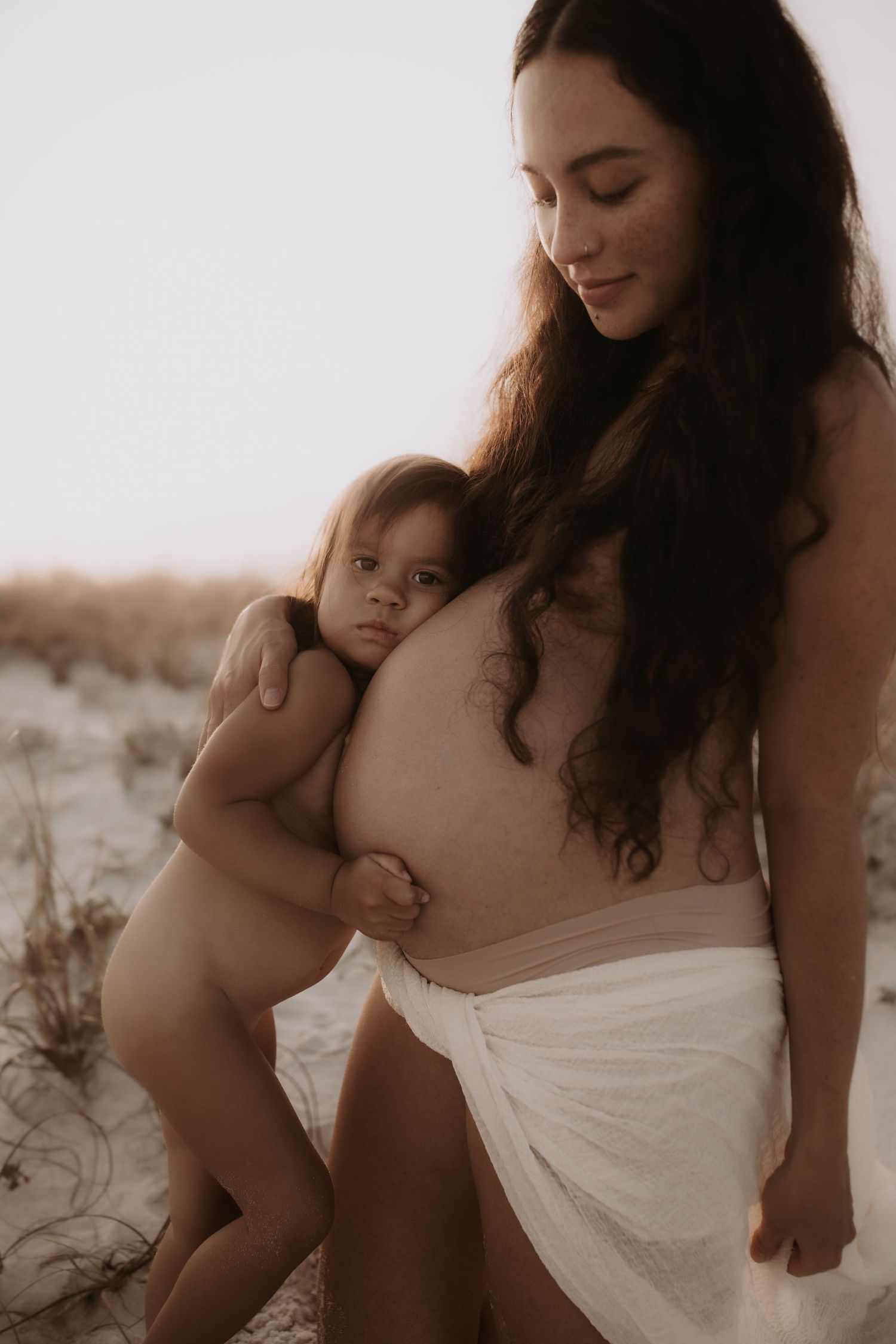 Smiling pregnant woman with long hair holds toddler on sandy dunes in soft maternity portrait.