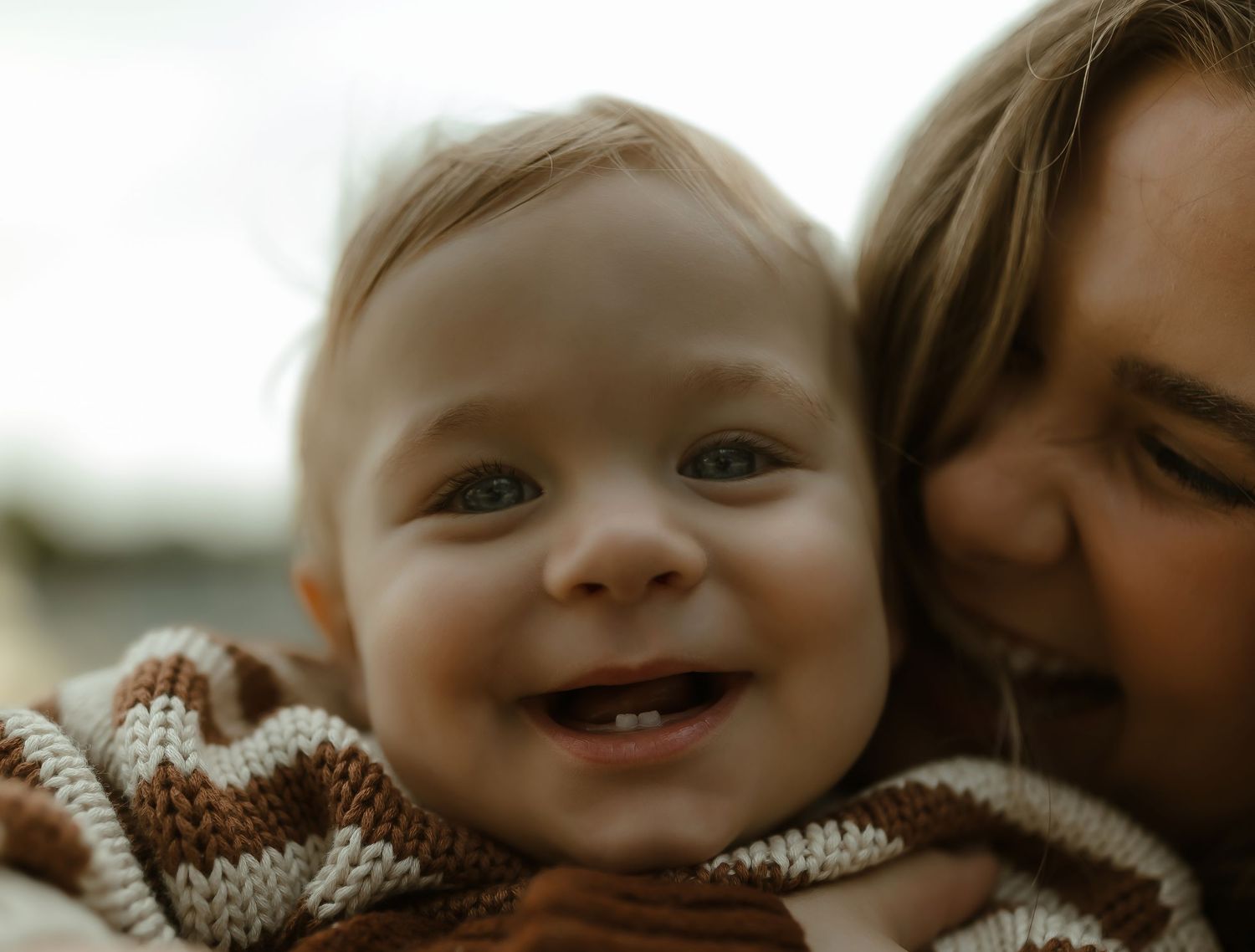 A joyful baby with blue eyes smiles brightly while being held by a woman in a cozy outdoor setting.