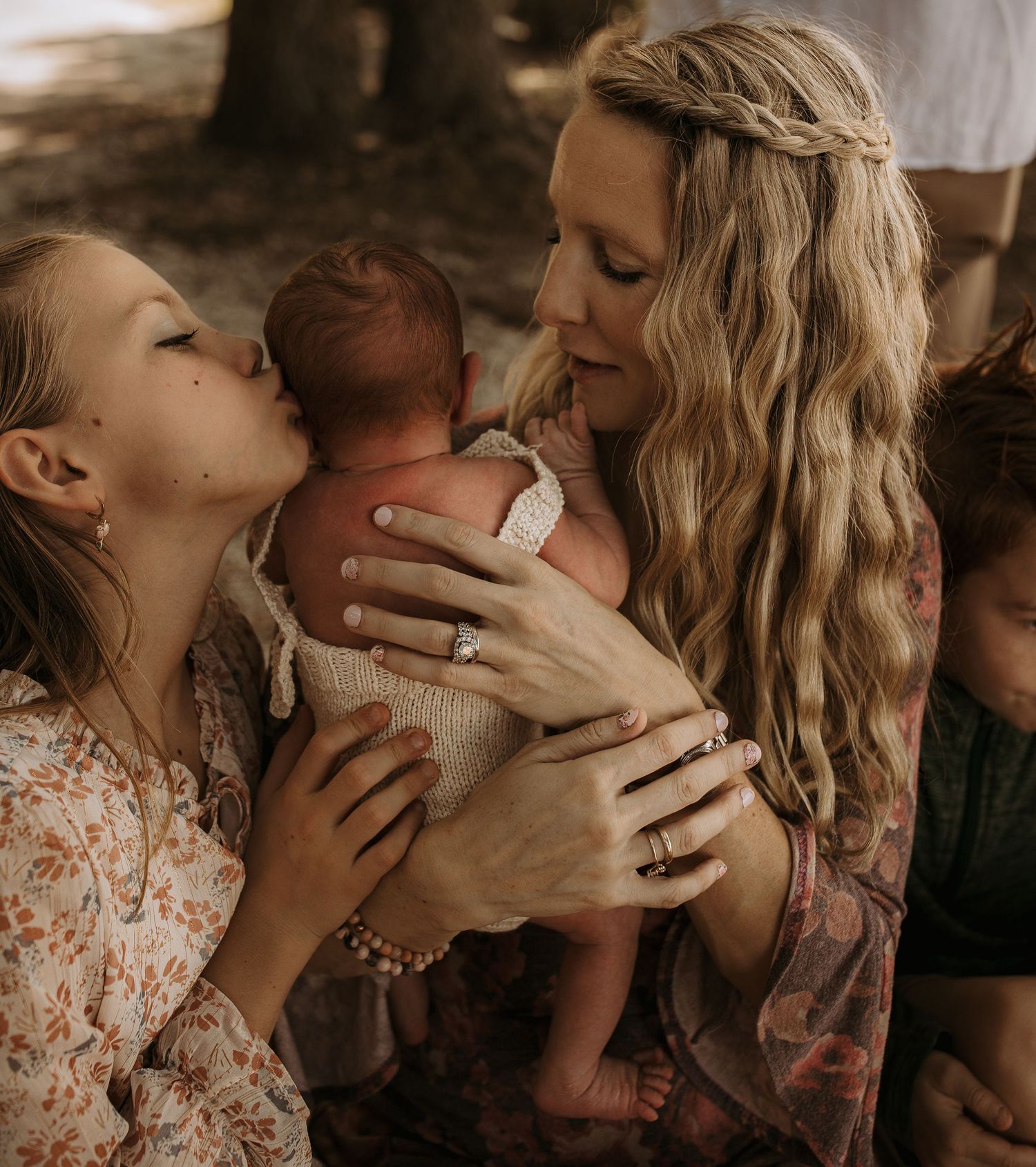 A blonde mother tenderly holds her newborn as an older daughter kisses the baby's head outdoors.