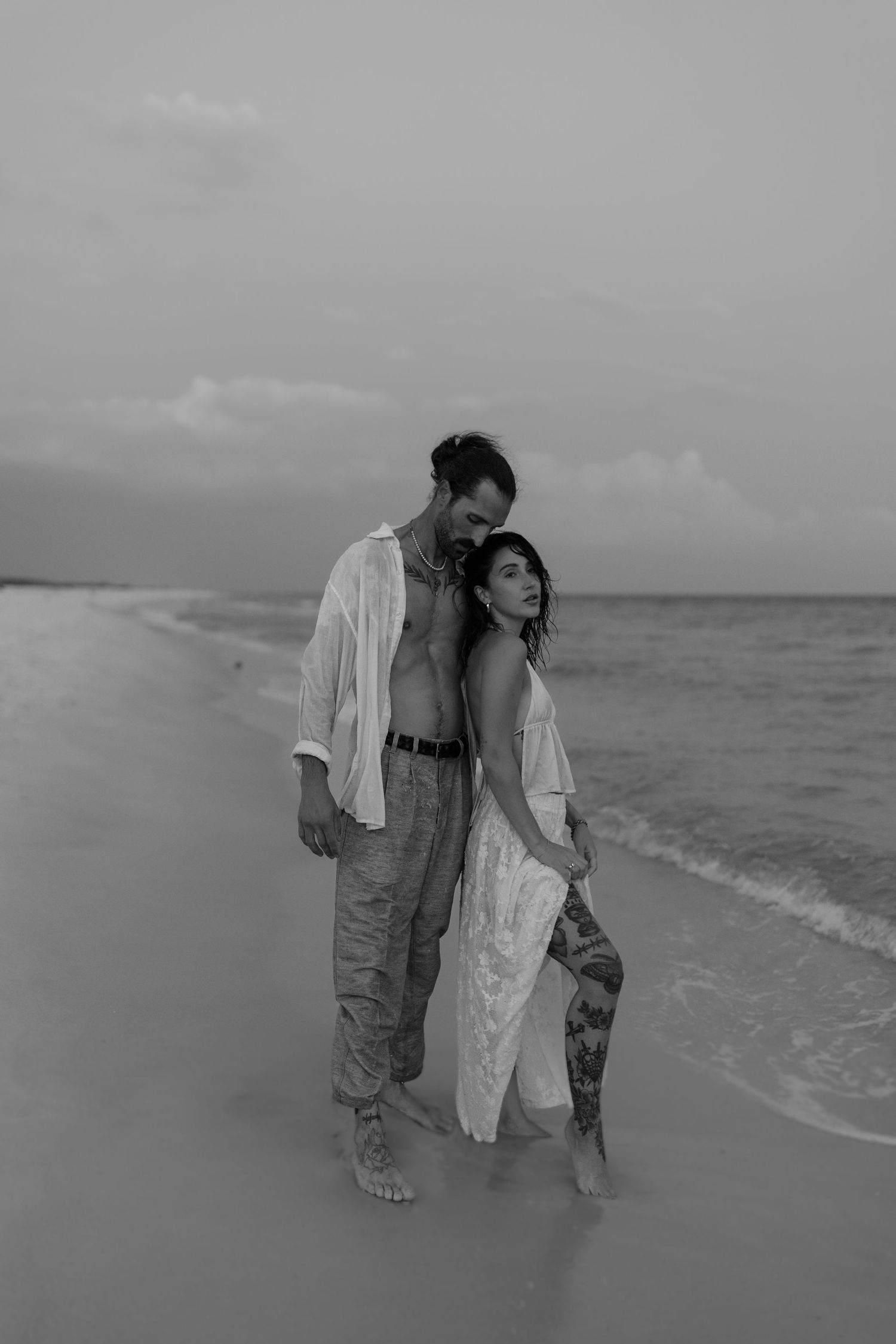 Black and white photo of a couple embracing barefoot on a sandy beach at dusk.