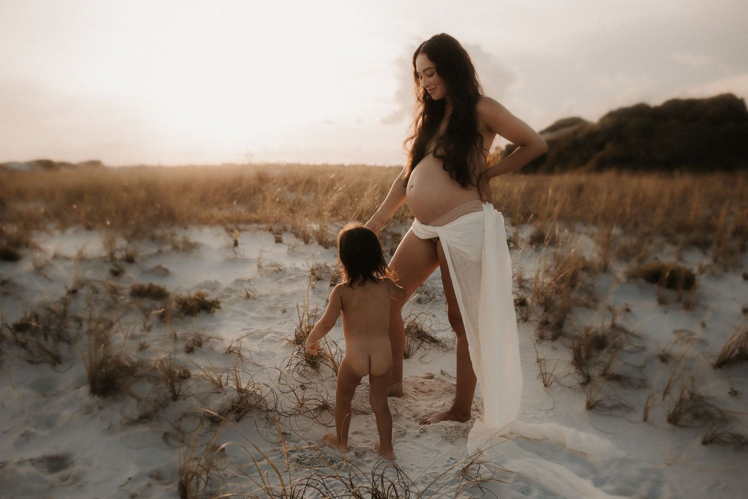 Pregnant woman in white fabric stands with toddler on white sand dunes at golden sunset.