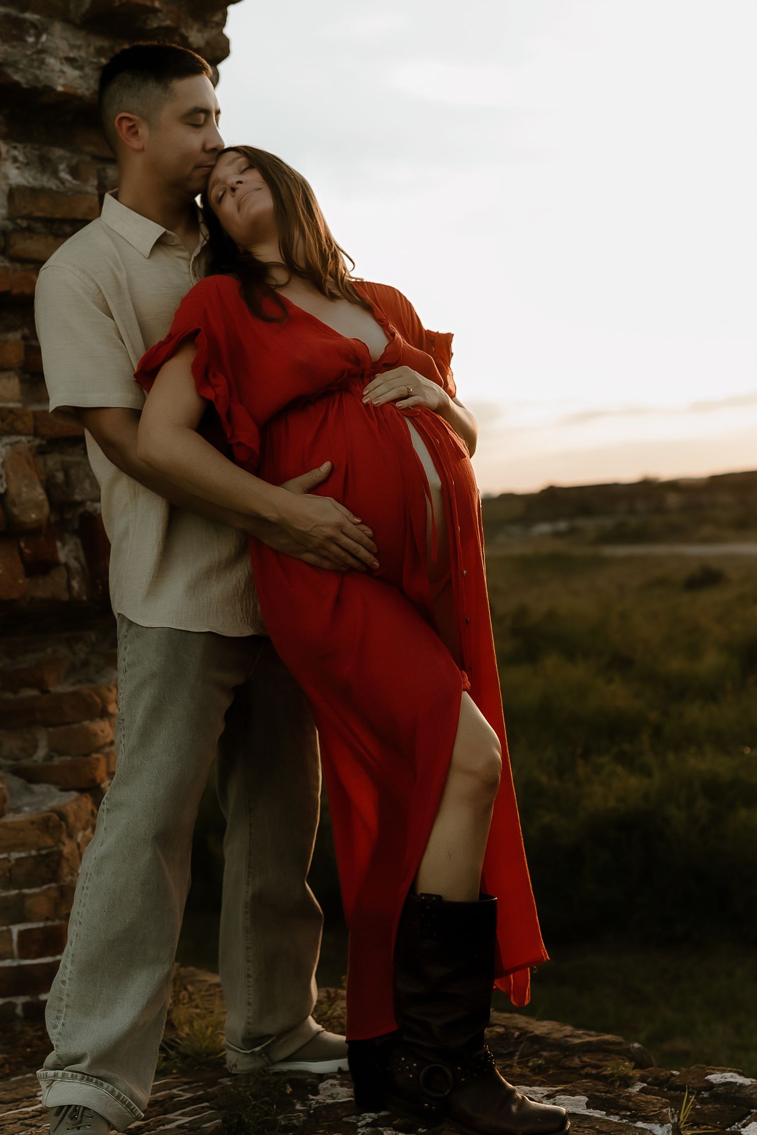 A couple embraces at sunset, the pregnant woman in a flowing red dress leaning into her partner.