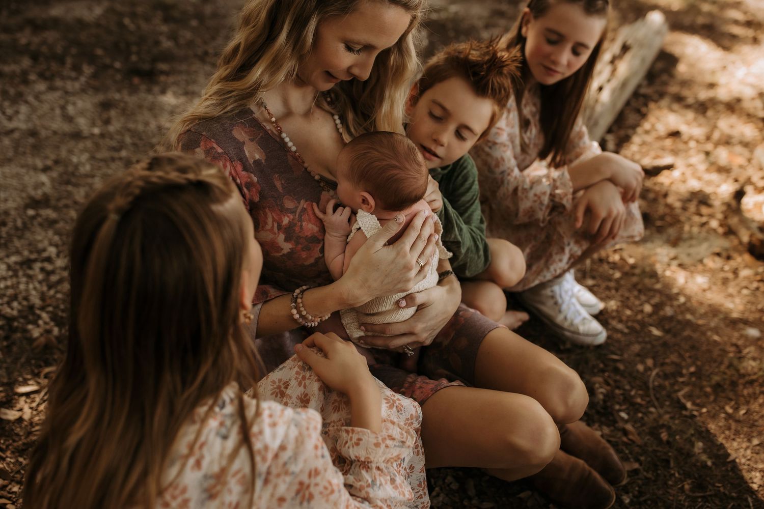 A mother sits cross-legged outdoors holding a newborn while surrounded by her daughters in floral dresses.