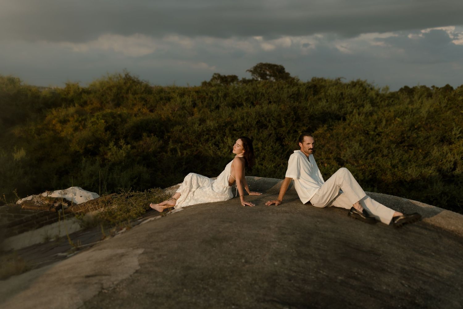 Couple in white outfits sitting on a large rock surrounded by lush greenery under a dramatic cloudy sky.