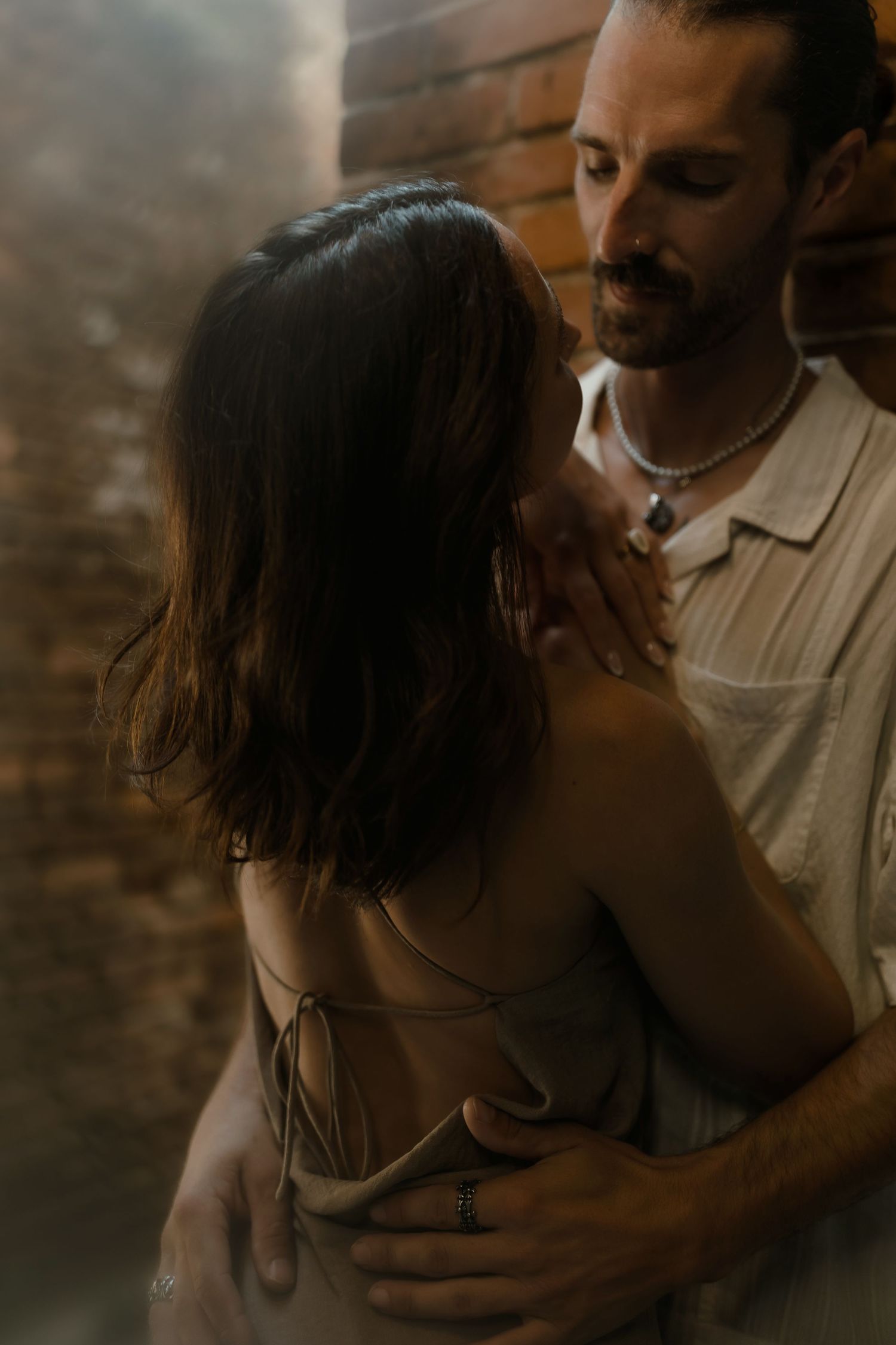 A bearded man in a linen shirt embraces a woman in a strappy dress near a brick wall in warm moody light.