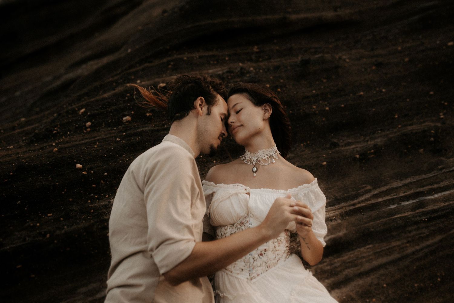 Romantic couple in wedding attire sharing an intimate moment against a dark rocky background.