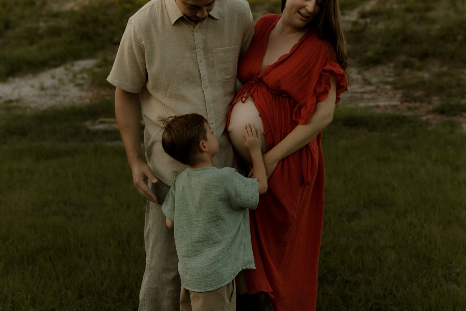 A family maternity photo with a young child touching the pregnant mother's belly outdoors in a green field.