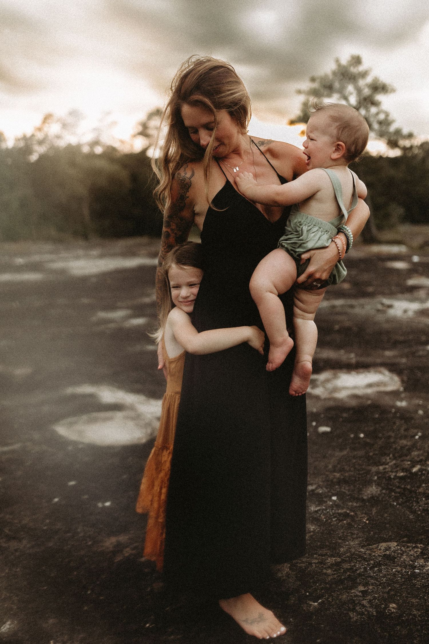 A mother in a black dress holds her baby while her young daughter hugs her at golden hour outdoors.