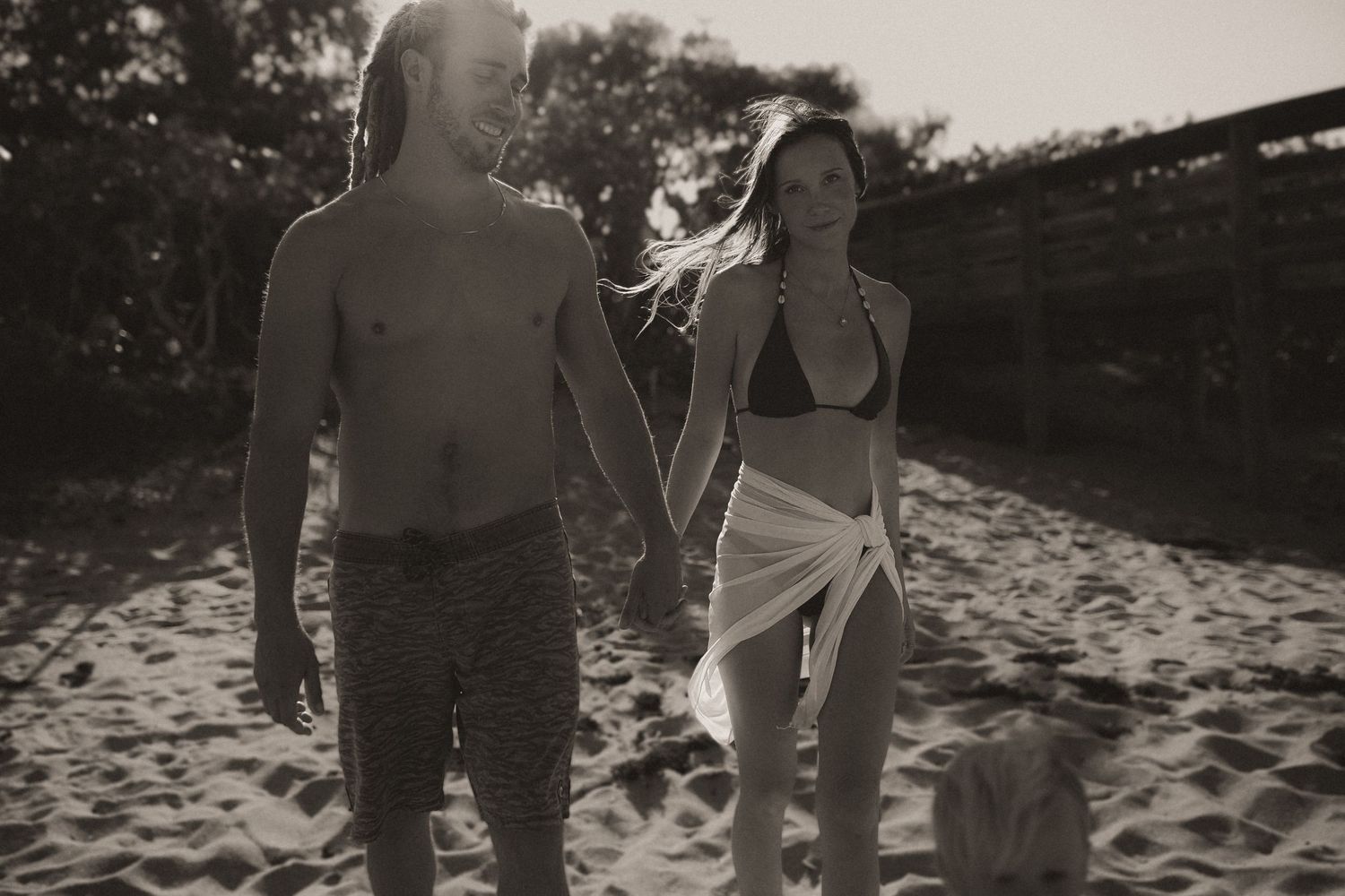 Couple holding hands walking on beach in black and white photo with golden backlight.