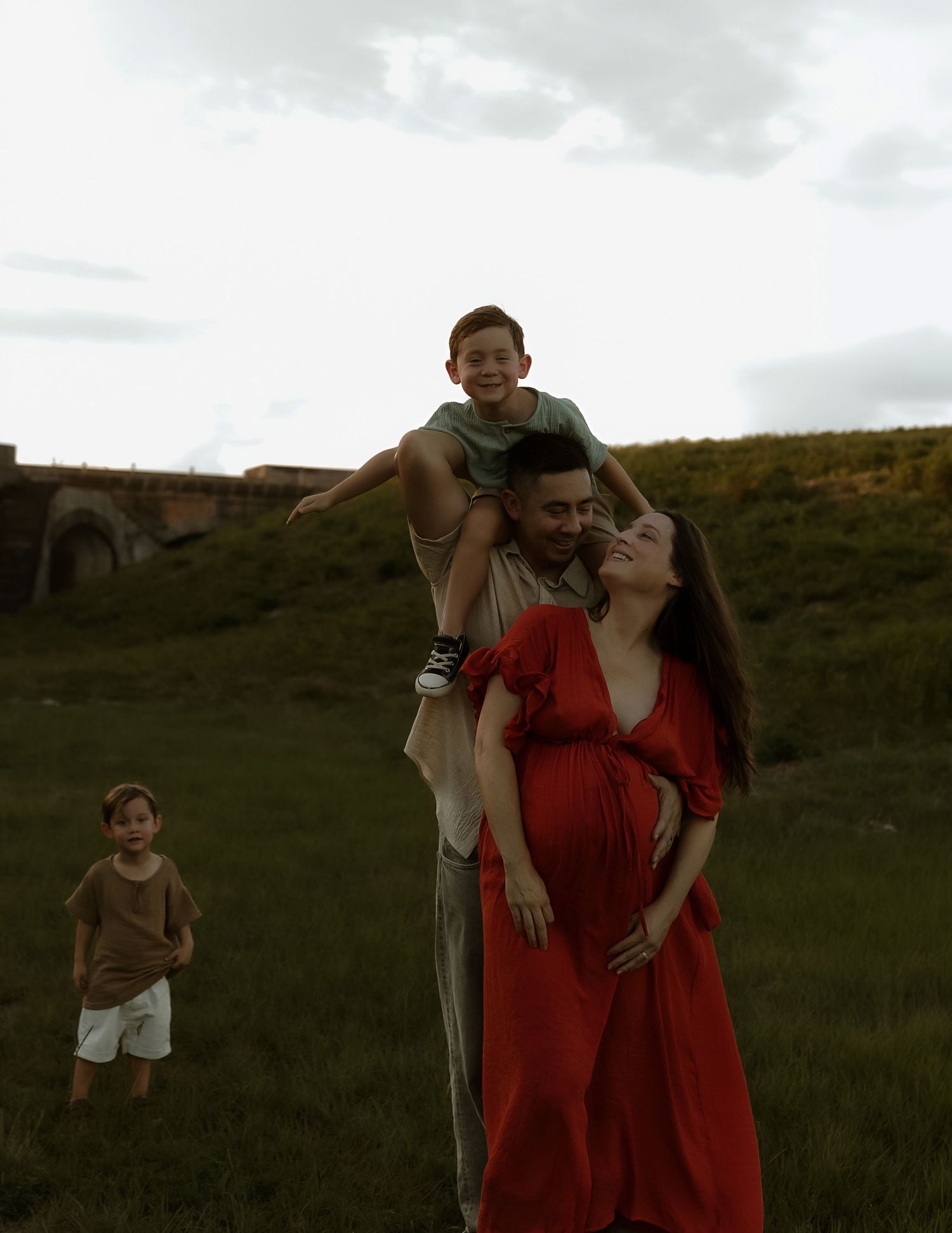 Pregnant woman in red dress with family outdoors on green field at dusk.