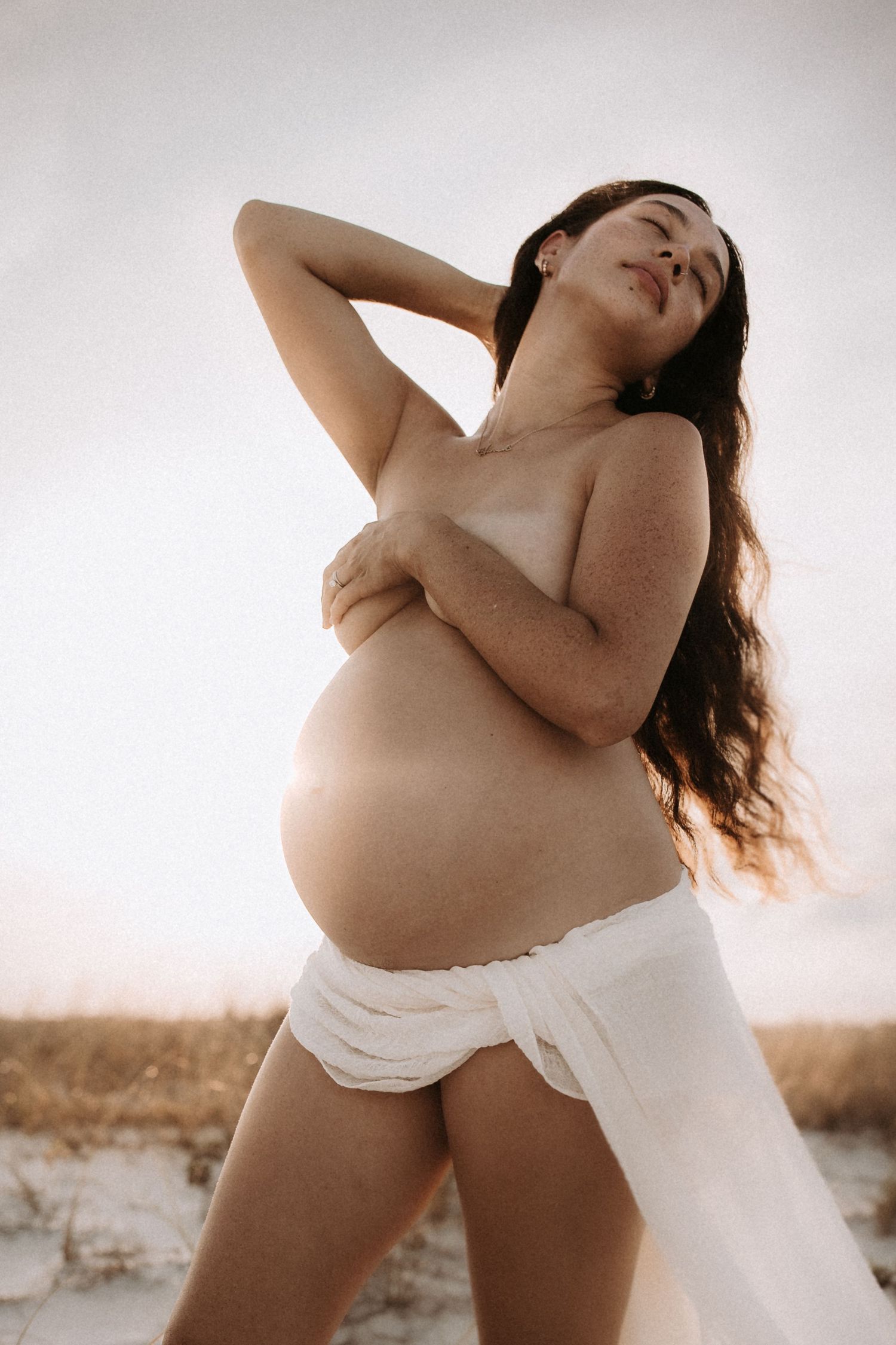 Pregnant woman posing outdoors at sunset, embracing her baby bump with flowing white fabric, natural maternity photography.