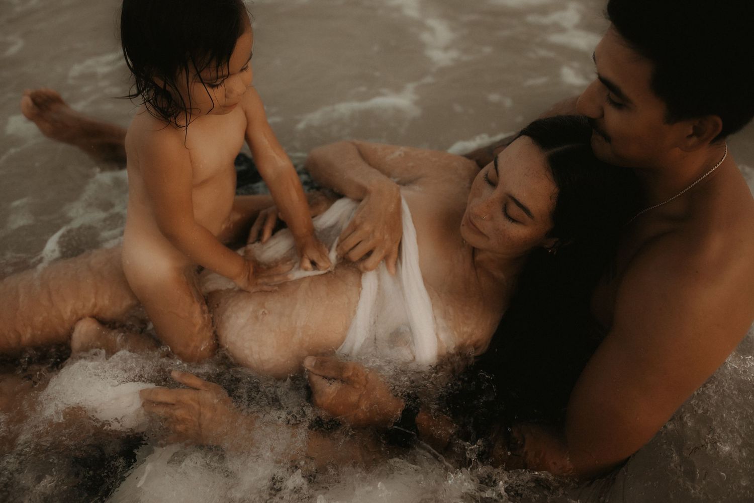 Family enjoying ocean waves together, with a mother in white swimwear holding her baby at the beach.