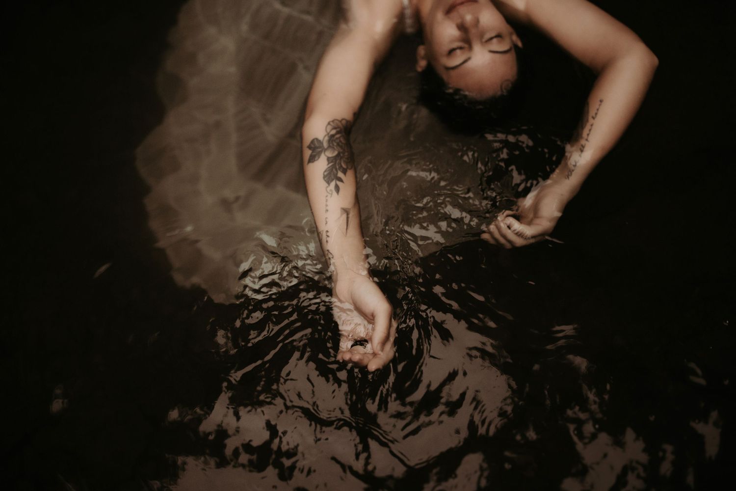 A tattooed woman floats in dark steamy water with eyes closed, wearing a flowing dress.