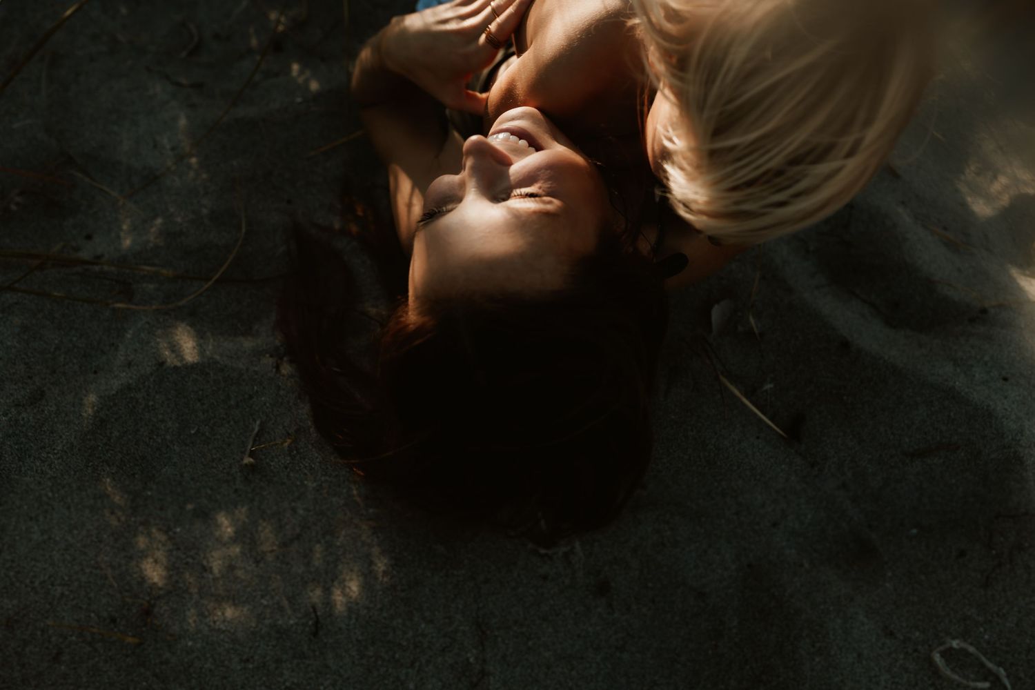 Overhead view of a woman with dark hair lying on sandy ground, smiling in soft natural light.