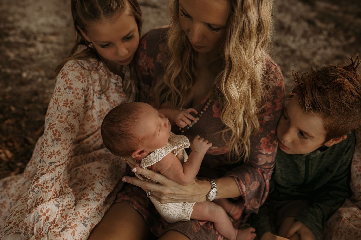 Mother with blonde hair holds newborn baby surrounded by two children in floral dresses outdoors.