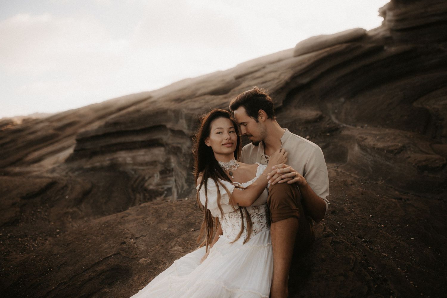 Couple embracing romantically on rocky terrain at sunset, woman in white dress, man in casual attire, outdoor engagement photo.