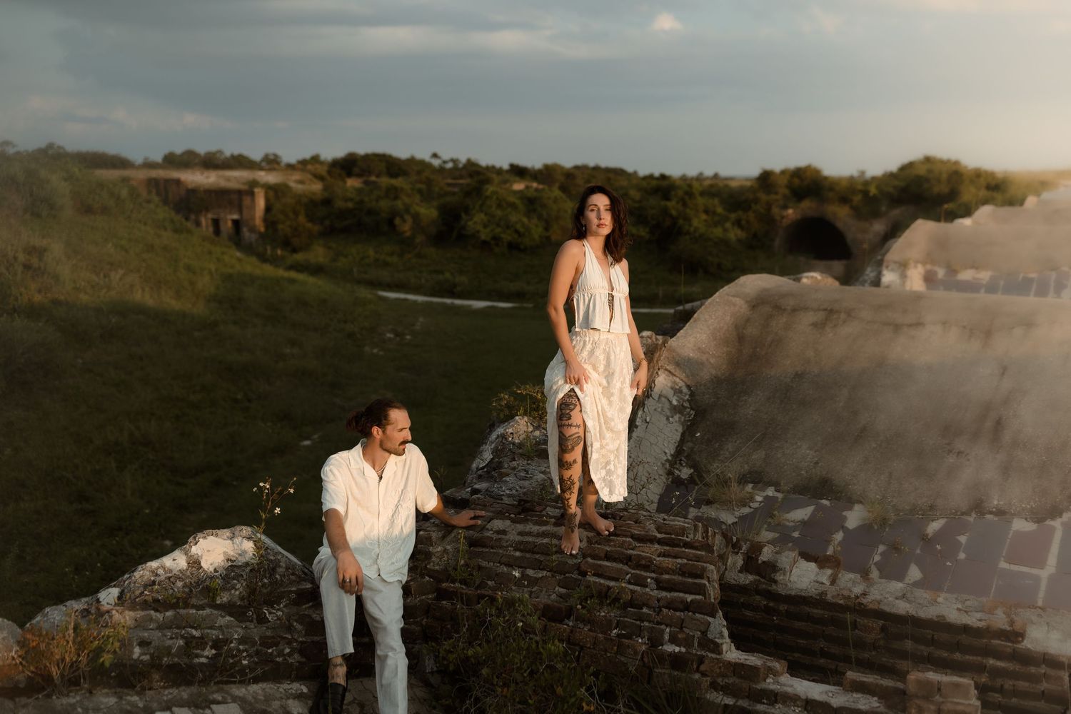 Couple standing on historic stone ruins surrounded by lush greenery at sunset.