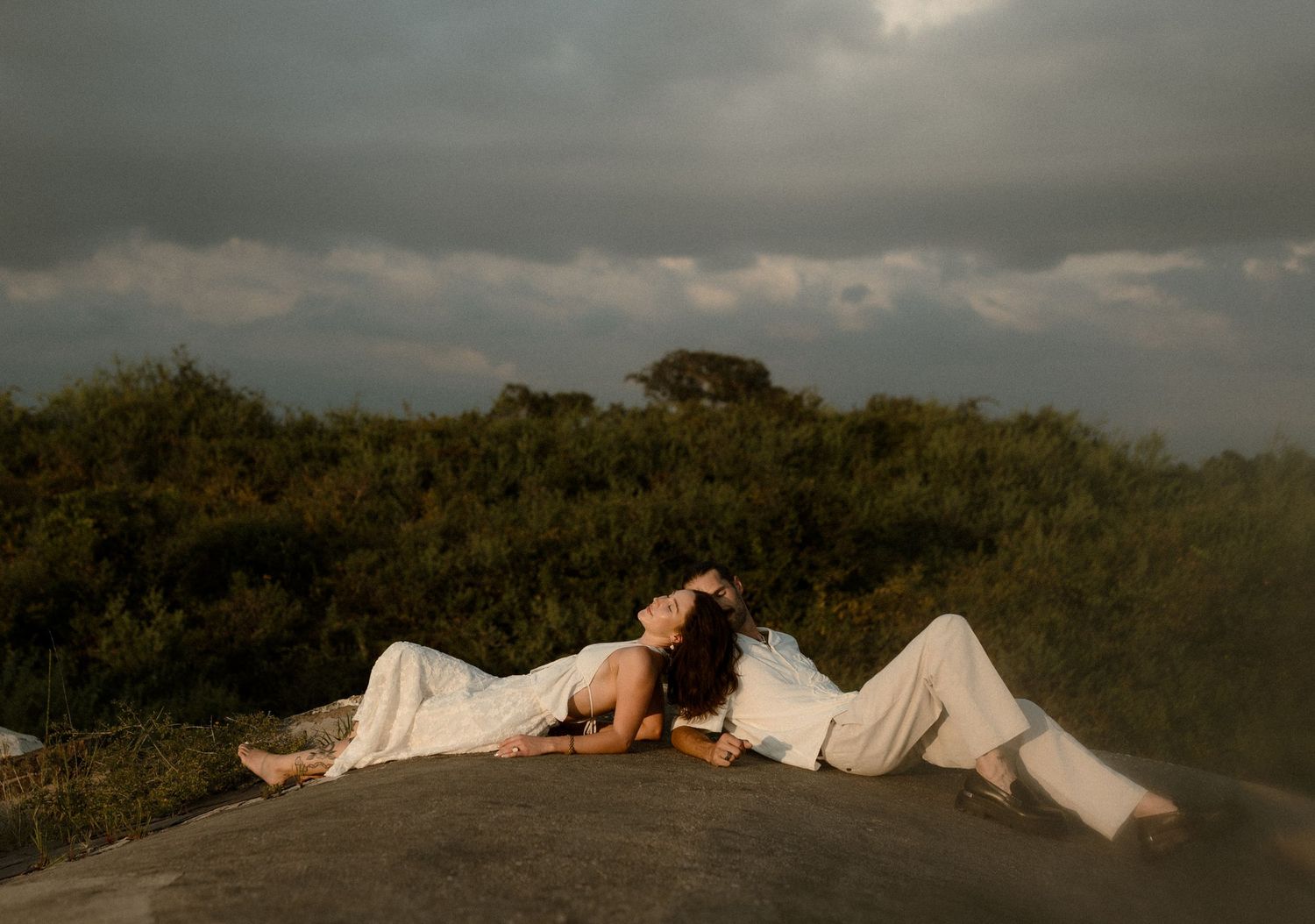 A woman in white clothing reclines gracefully on a rock with lush greenery and dramatic cloudy sky behind her.