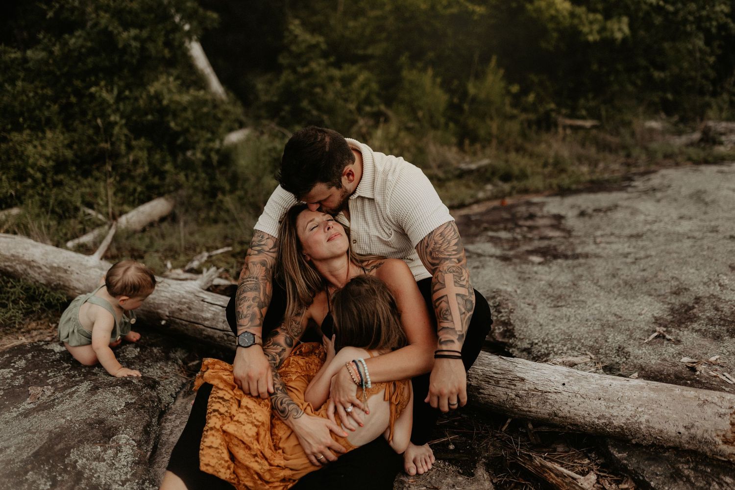 Tattooed couple embracing children near a fallen log in a natural outdoor family portrait session.