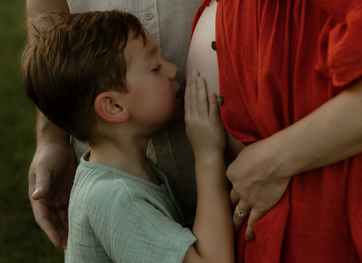 Young boy kissing pregnant mother's belly outdoors, wearing light green shirt, warm family maternity photo.