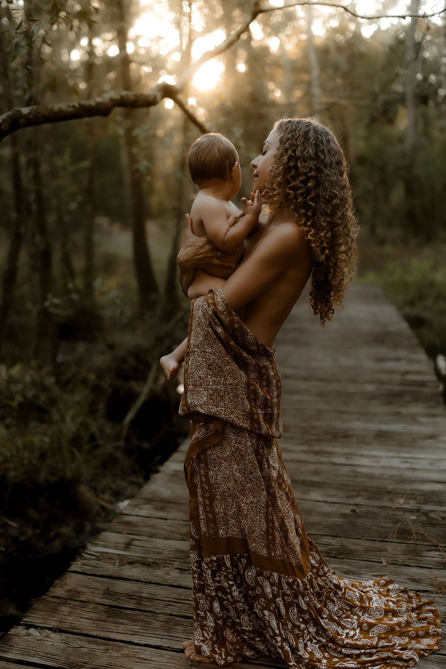 Mother lifts laughing baby overhead on woodland boardwalk bathed in golden sunset light.
