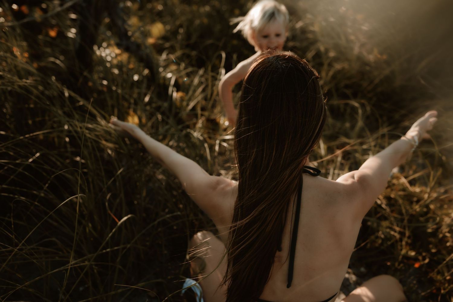 A mother with long hair sits in sunlit grass with arms outstretched as her toddler stands behind.