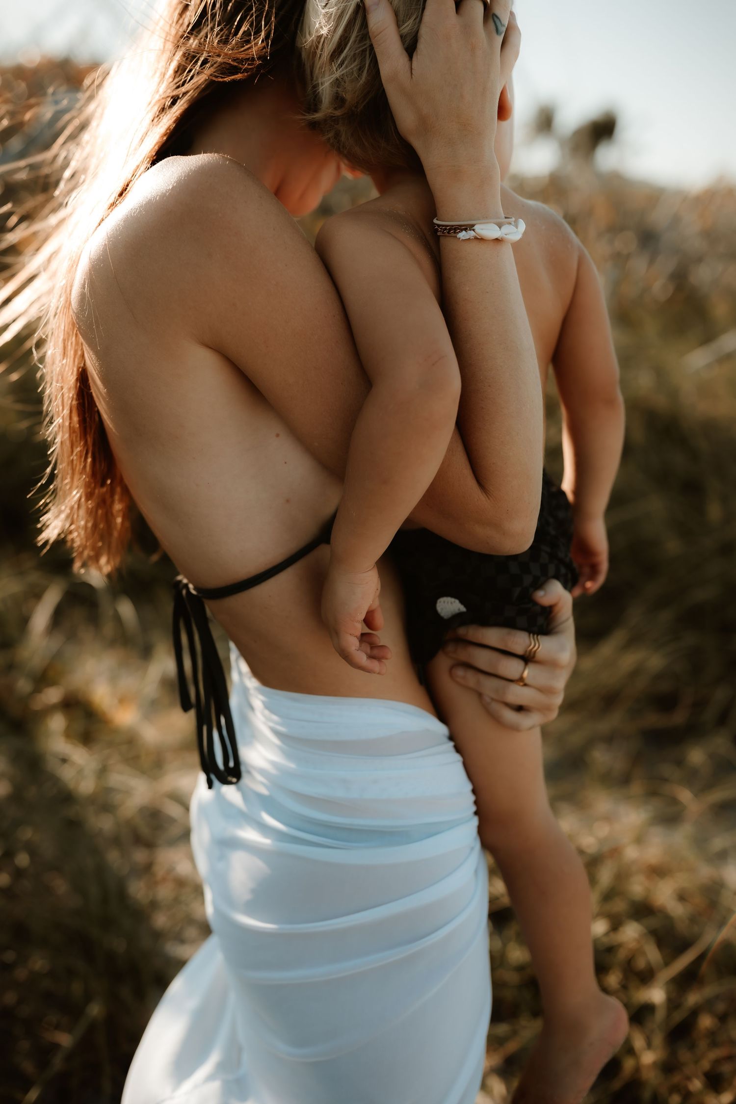 A mother in a black bikini top and white skirt holds her baby outdoors in golden grass.