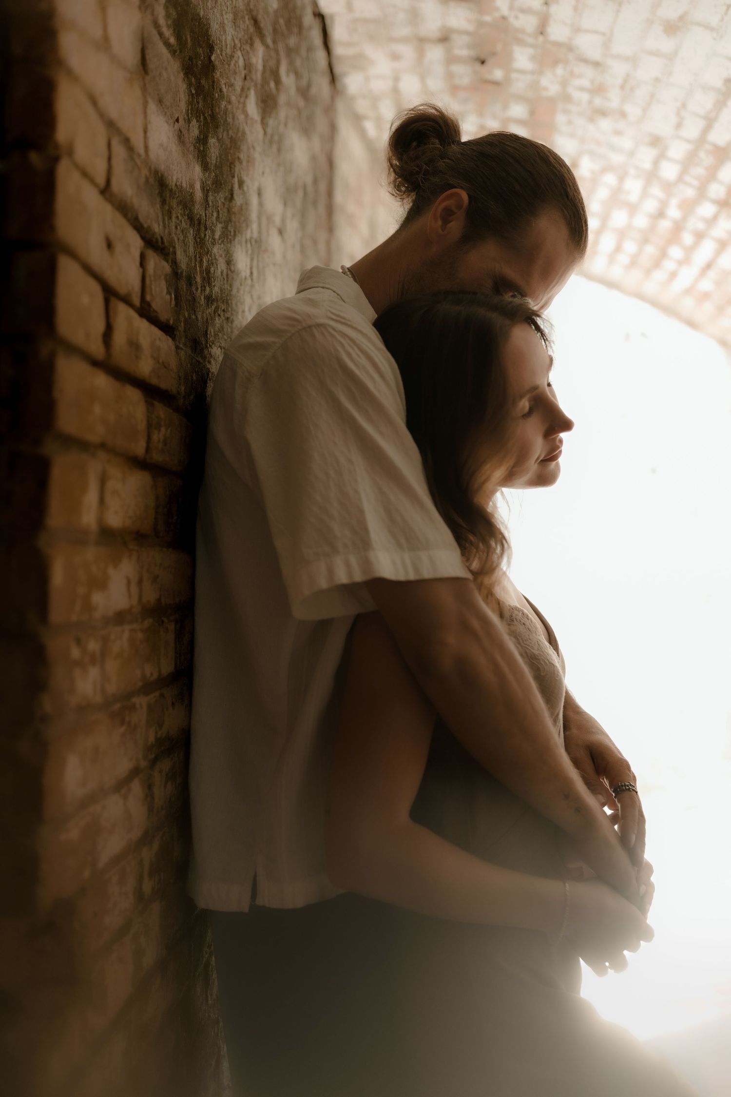 A couple embraces tenderly against a rustic brick wall in soft warm light, the man holding the woman from behind.