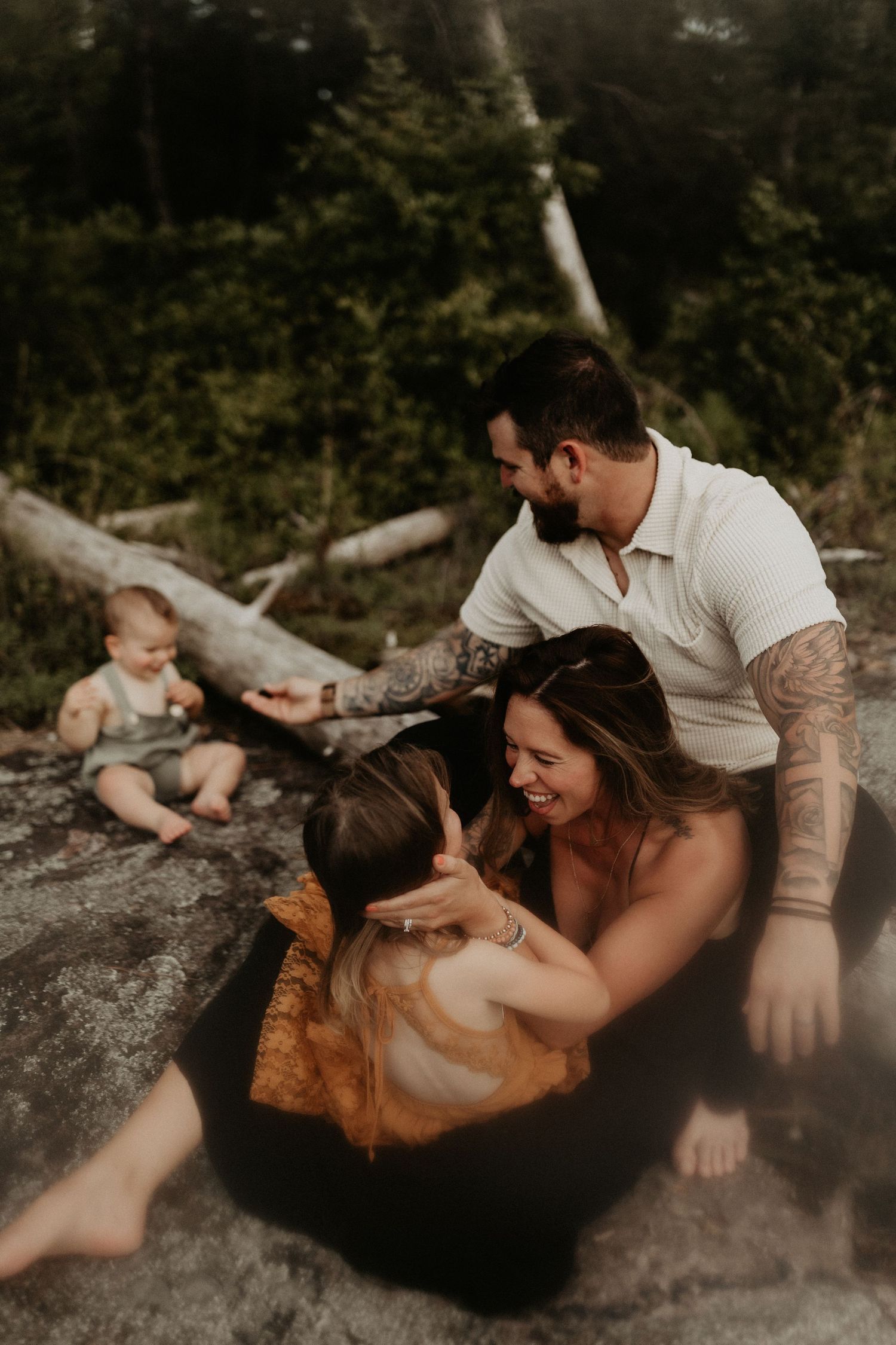 Tattooed father and smiling mother sit on rocks outdoors with two young children in a candid family portrait.