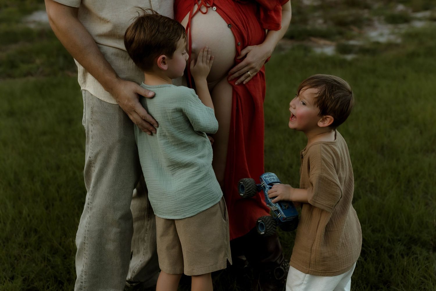 Two young boys touching a pregnant womans belly outdoors during a maternity photo session in a green field.