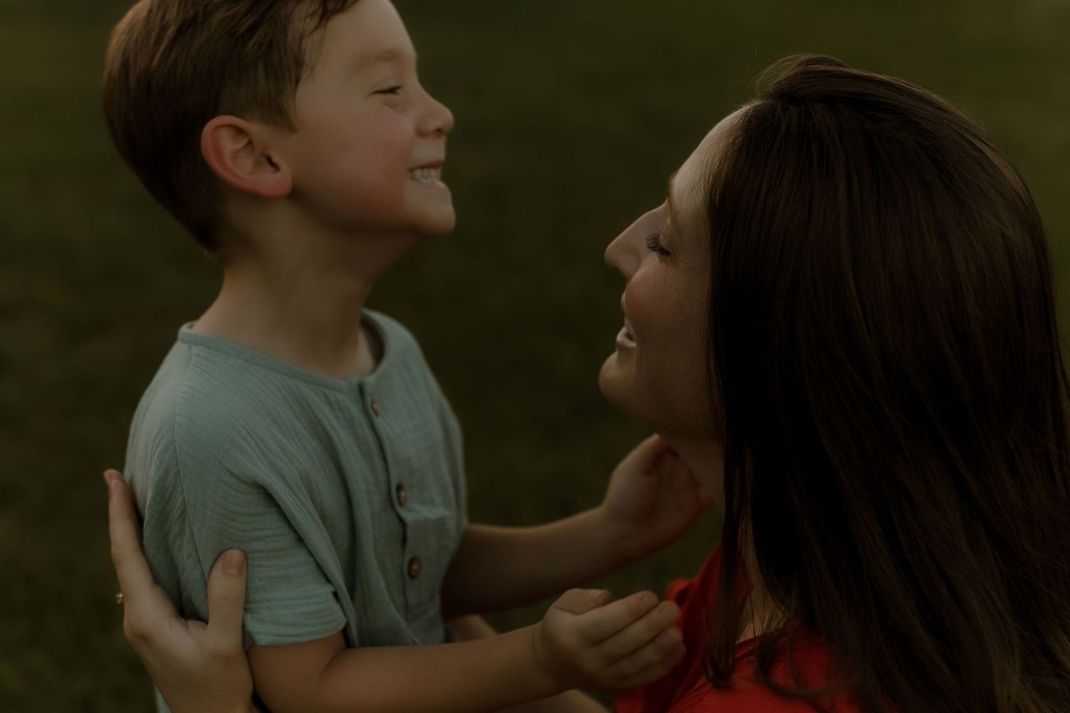 A young boy and woman share a joyful moment outdoors, smiling at each other in a warm embrace.