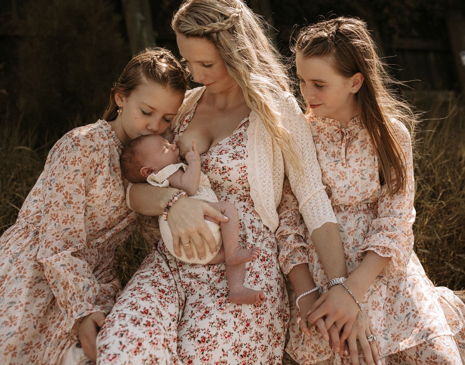 A mother in floral dress breastfeeds newborn baby while two daughters in matching dresses admire the infant outdoors.