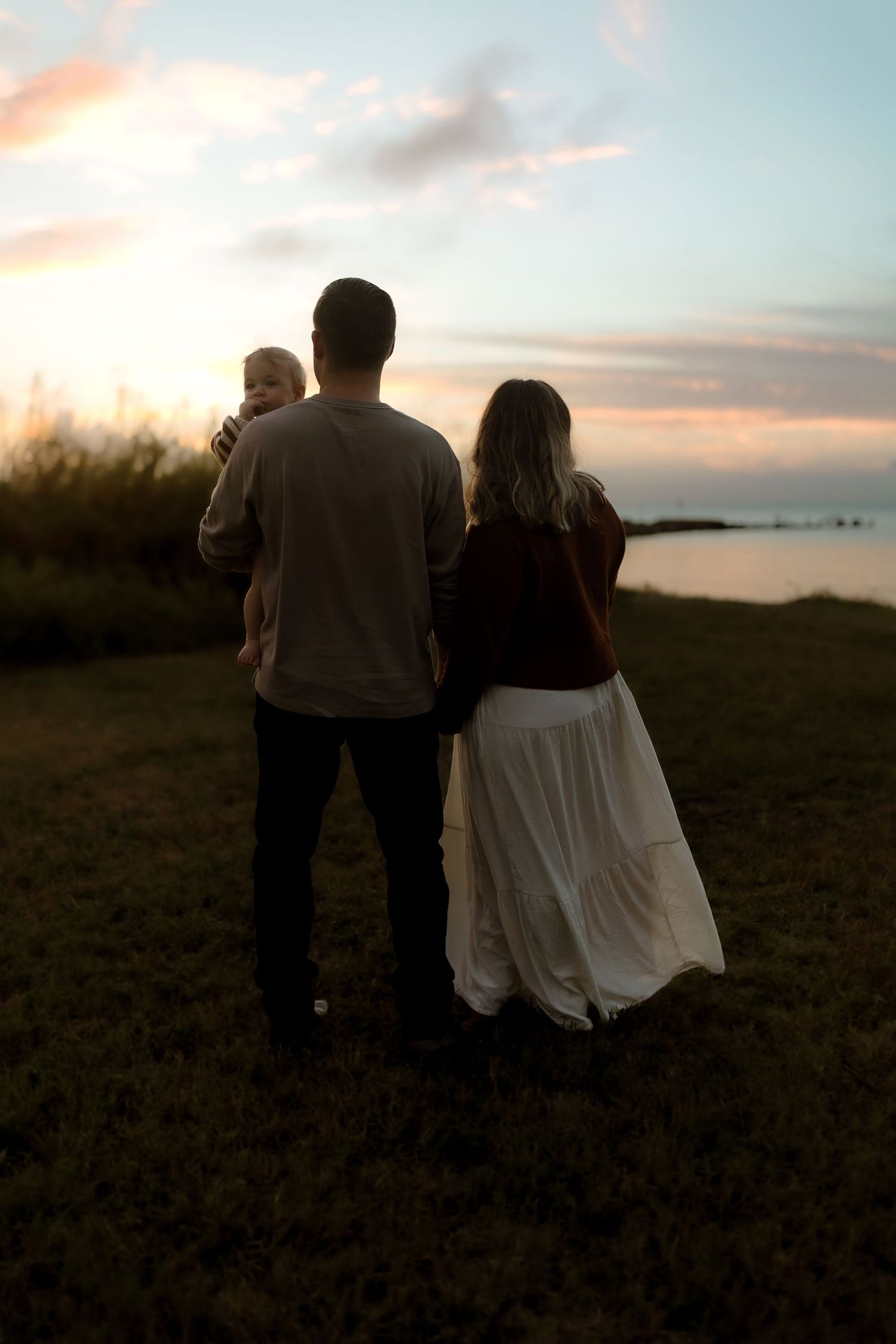 Family of three standing on grass at sunset, parents holding baby while gazing at colorful sky over water.