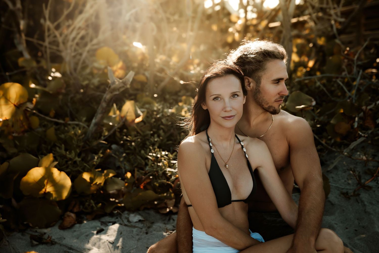 Woman in black bikini top sitting with shirtless man in golden sunset beach setting.