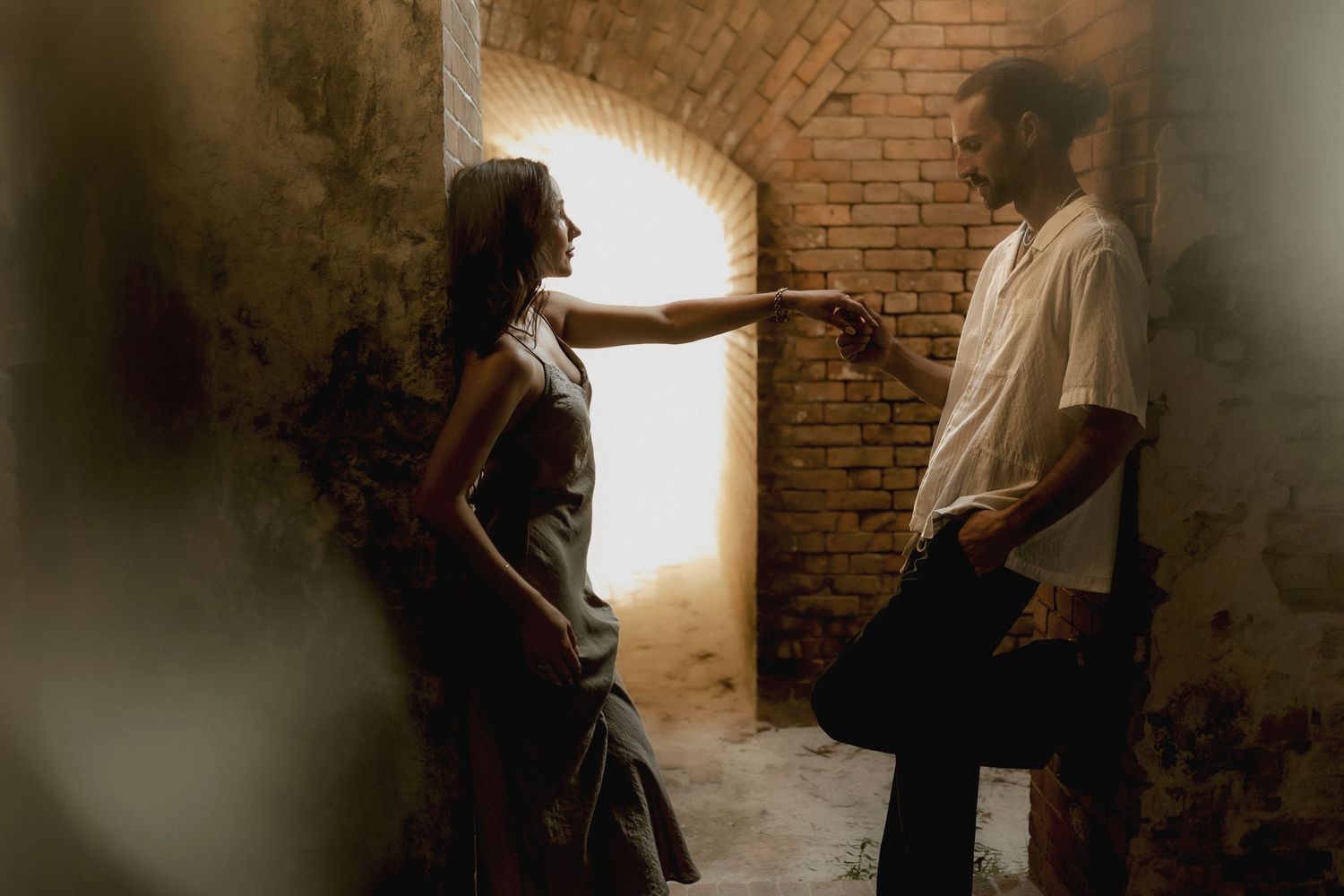 Two people face each other in a dimly lit brick tunnel, one extending a hand toward the other near a glowing arched doorway.